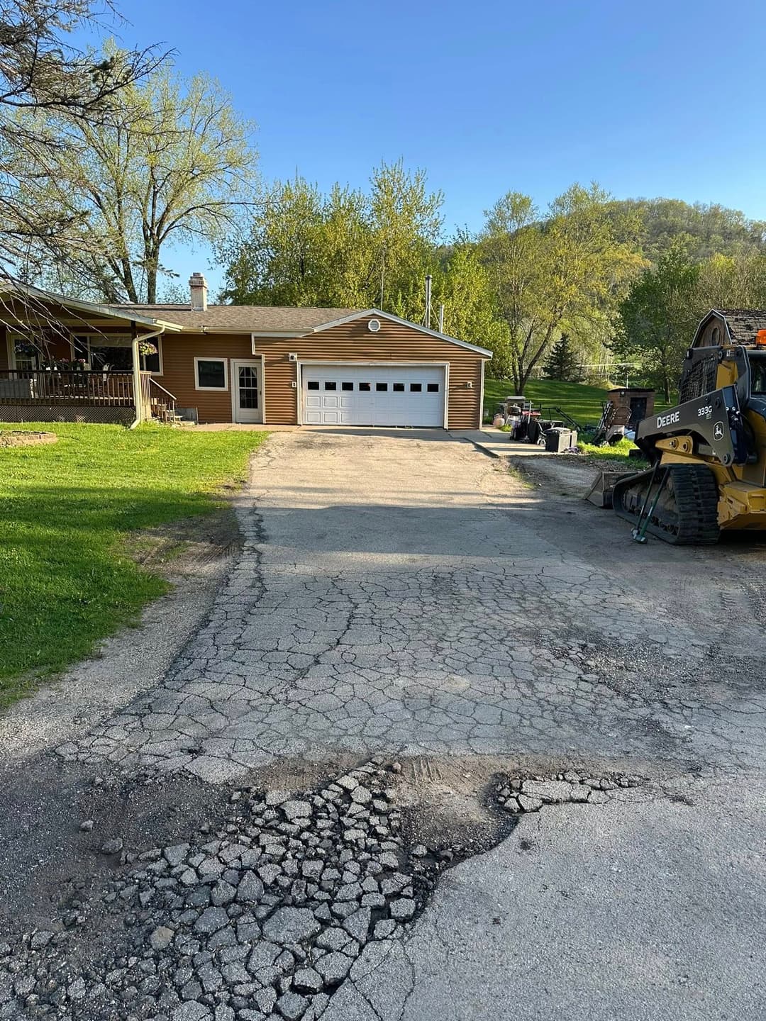 Residential home with a garage, cracked driveway, and construction equipment nearby in a green landscape.