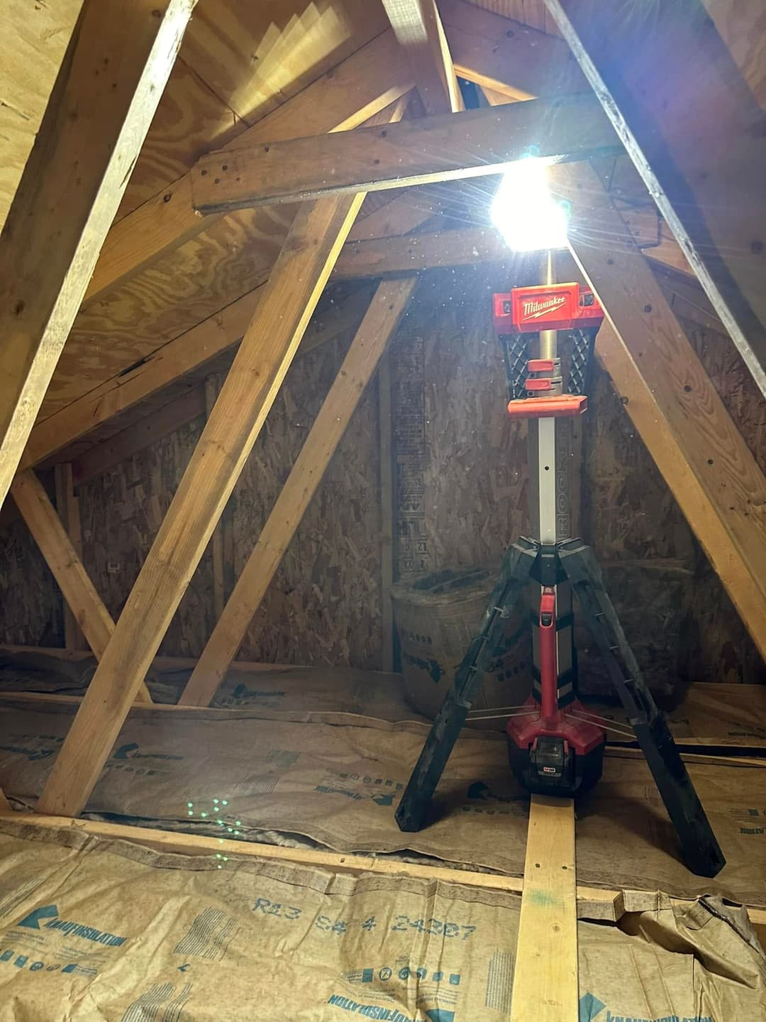 Bright work light on tripod illuminating a wooden attic space with exposed beams and floor covering.