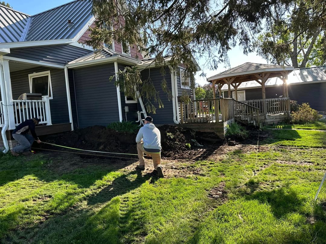 Workers measuring and preparing landscaping near a house and gazebo in bright sunlight.