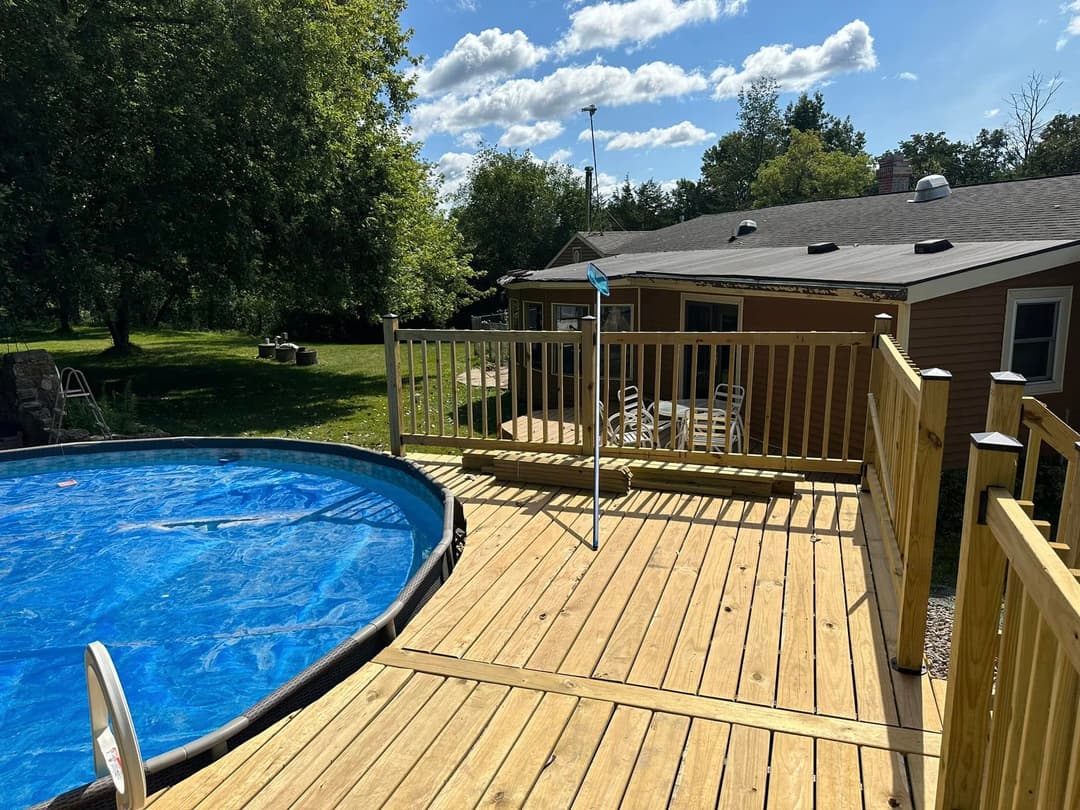 Wooden deck with an above-ground pool, surrounded by greenery and a house in the background.