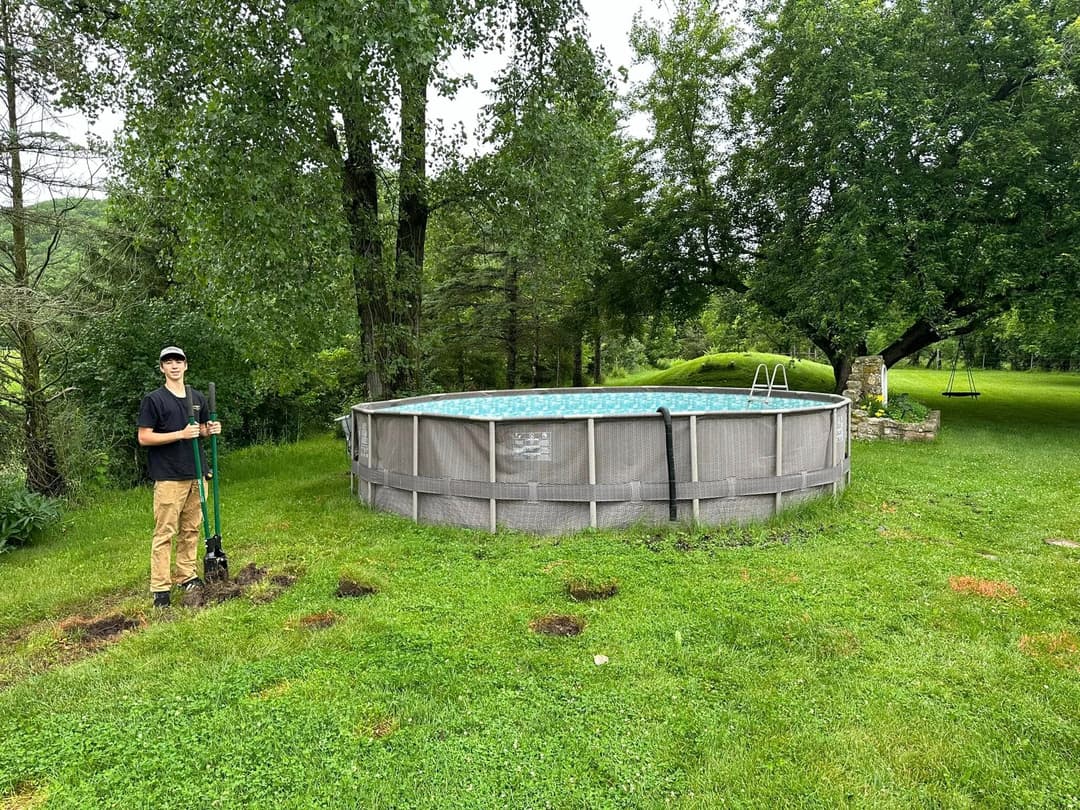 Teenager standing with a shovel next to an above-ground pool in a grassy backyard.