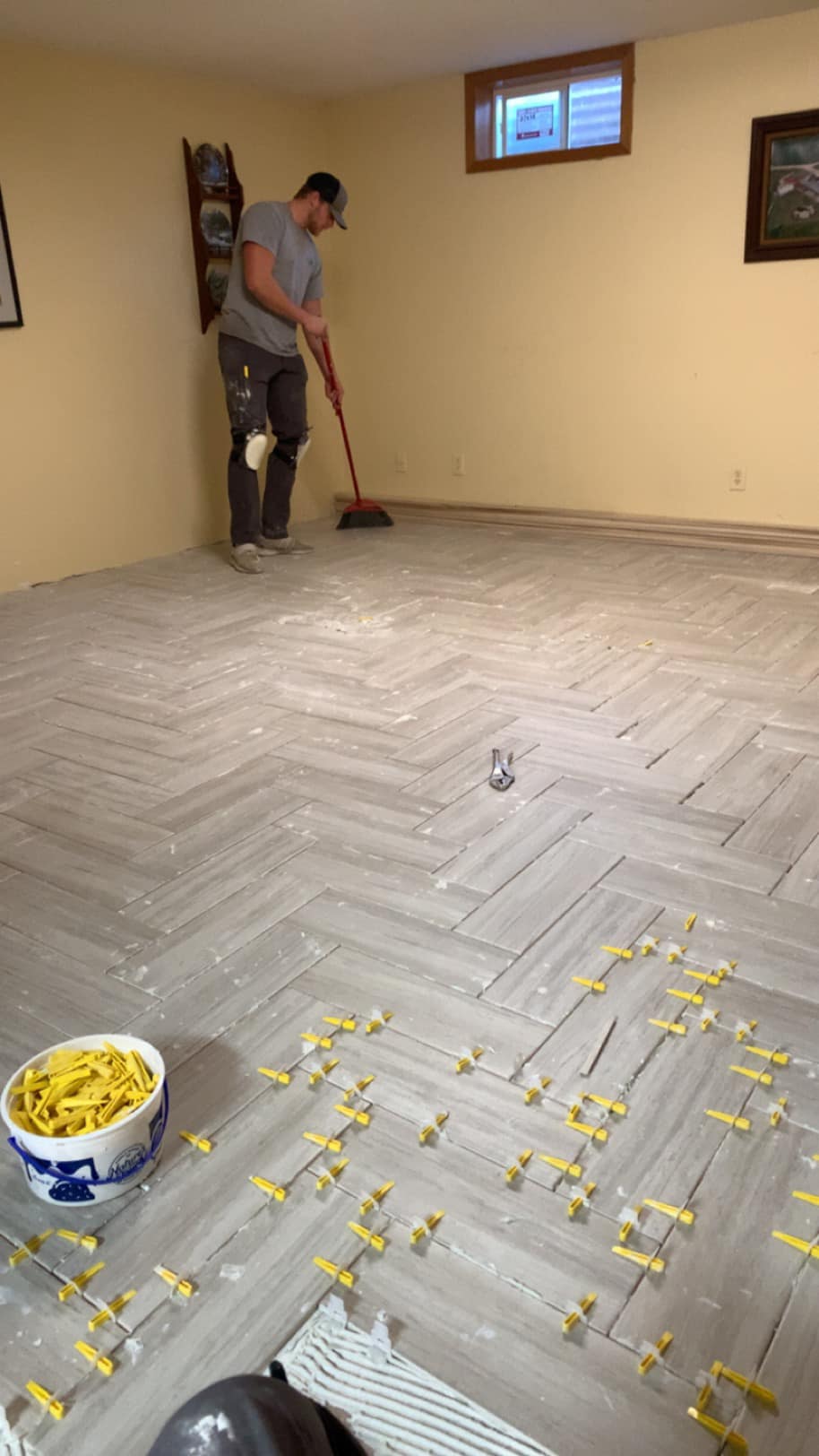 Man sweeping a newly tiled floor with tile spacers and tools in a basement room.