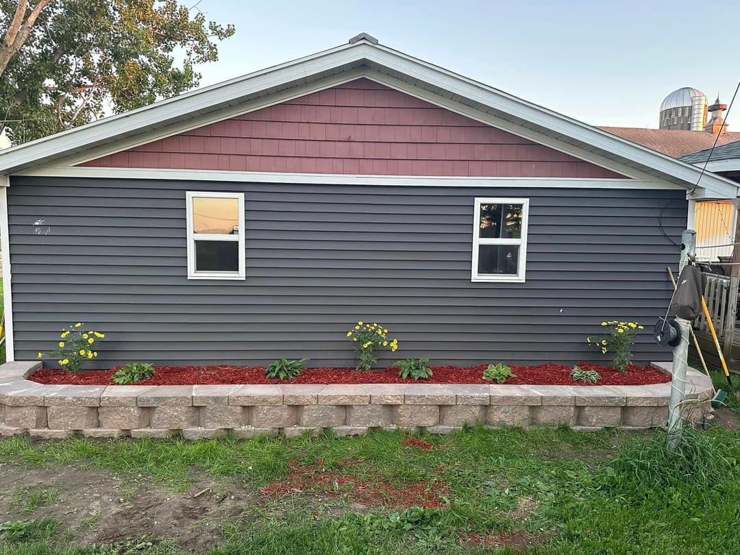Back of a house with gray siding, maroon accents, and a flower bed featuring yellow flowers.