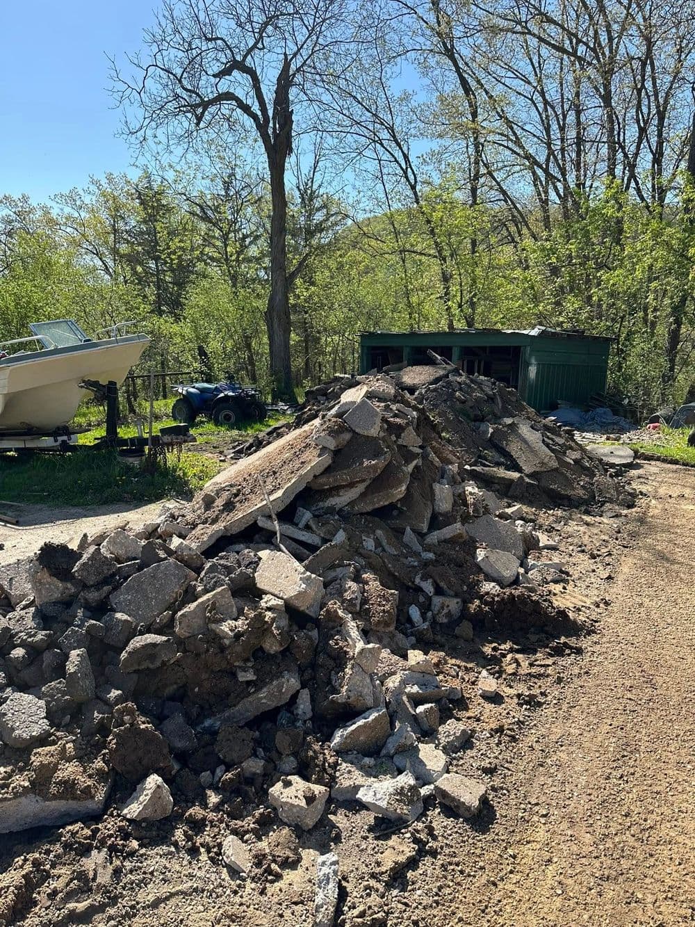 Piled concrete debris near a shed with a boat and ATV in a lush green landscape.