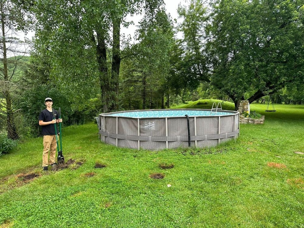 Teenager standing with a shovel beside an above-ground swimming pool in a green backyard.