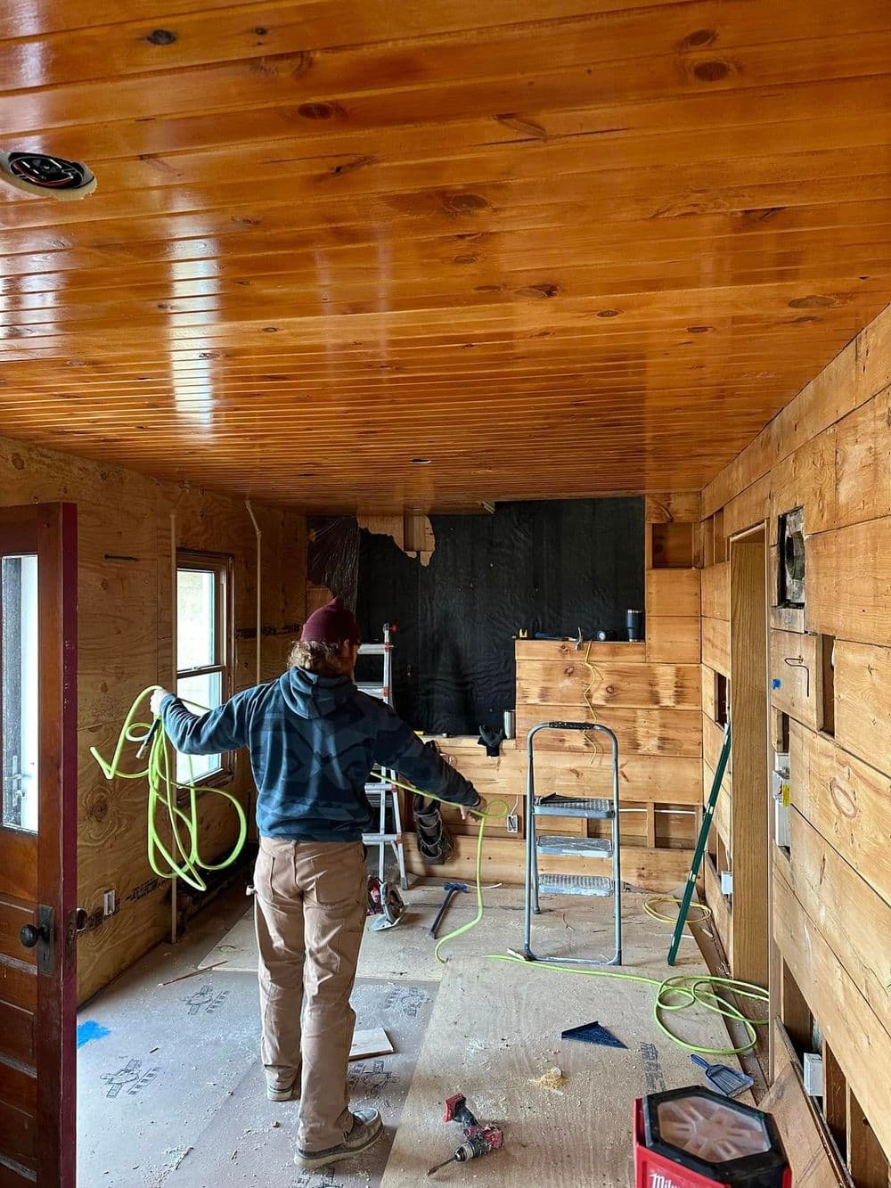 Person renovating a wooden ceiling in a rustic cabin interior with construction tools visible.